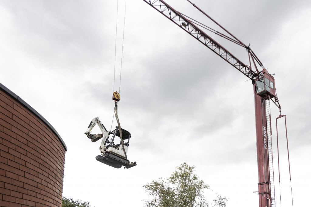 Ein Gärtner Bagger wird mit einem Kran über ein Haus hochgehoben, wegen schlechter zugänglichkeit